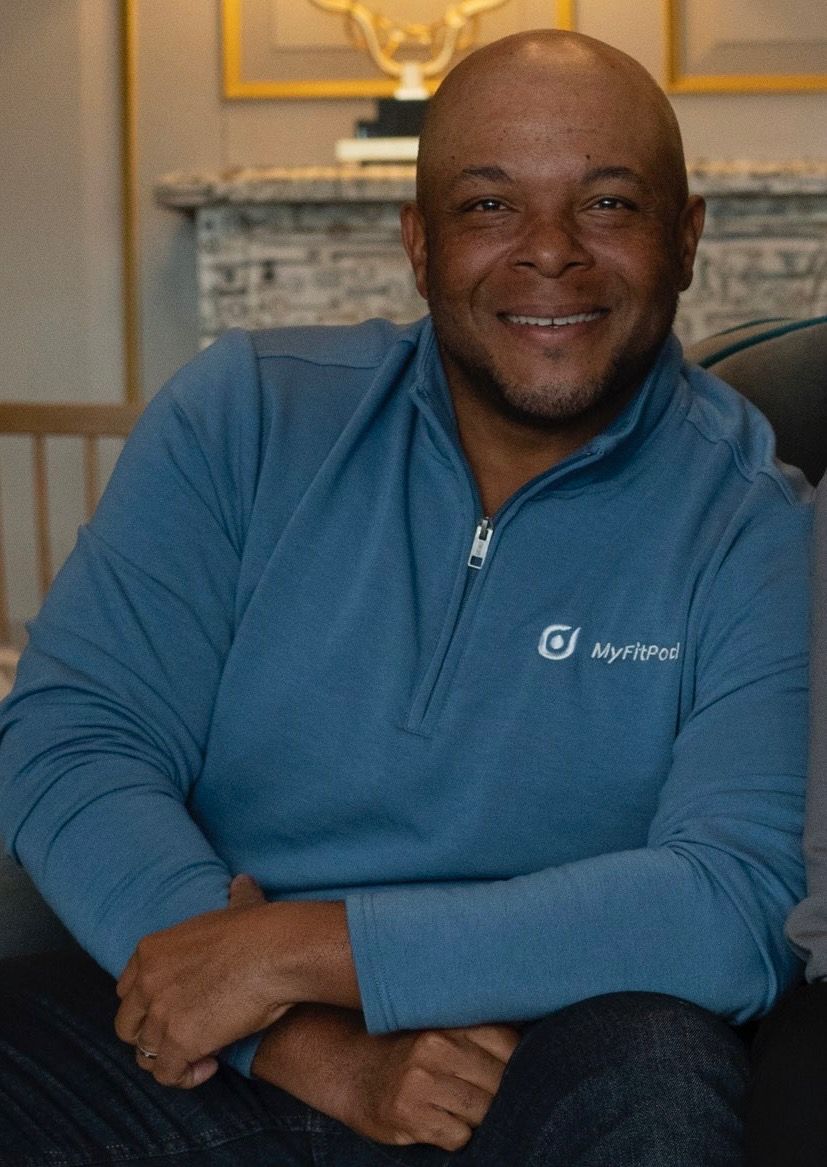 Rodney J. Morris, a bald, smiling black man, with in a chair with his wrists crossed on his lap. He wears a blue zipped collared shirt with the words MyFitPod embroidered on his left side in white thread.